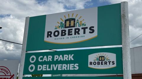 A turquoise-green sign with "Roberts: Raised in Cheshire" and car park and deliveries directions written on it