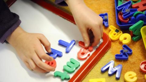 A children's hands organising magnetic letters, which are green, blue and red, on a whiteboard. Other letters surround the board on a yellow mat.