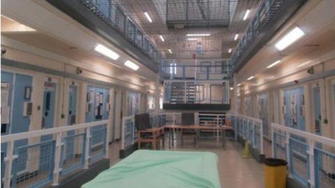 A view of the prison landing on a wing of HMP Risley, with blue cell doors surrounding a central area which has tables and chairs and a pool table with a light green covering. 