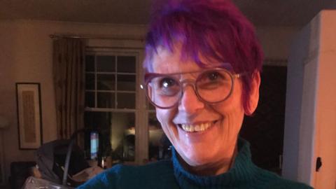 A woman with spiky purple hair and glasses, wearing a blue roll neck, smiles while stood inside a dimly-lit living room.