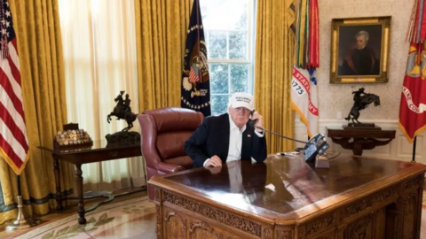 Donald Trump wearing a blue suit, white shirt and a white MAGA cap, sitting behind the Resolute Desk in the Oval Office holding a telephone to his ear