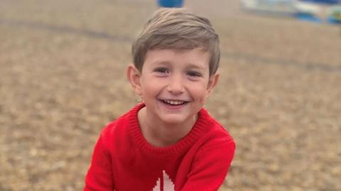 Young boy, Benedict, wearing a bright red jumper looks towards the camera outdoors, with a softly blurred background creating a natural portrait.