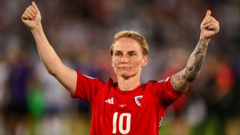Jess Fishlock in the Wales number 10 jersey holds her arms in the air with thumbs up to the crowd