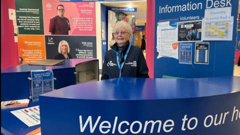 Betty Miller standing at the information desk in the lobby of Coventry's University Hospital. She has white hair and is wearing her blue volunteer's uniform with a volunteer lanyard. She is standing in front of a blue sign marked "Information Desk".