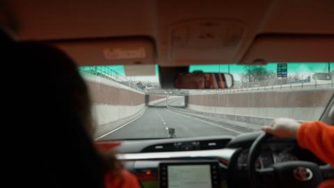 The view from inside a vehicle as it travels down a newly built underpass, with the road curving gently ahead and concrete walls on both sides. The dashboard and steering wheel are visible in the foreground, along with the reflection of the driver’s face in the rear‑view mirror.