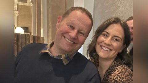 Jason is a bald man with blue eyes and he is smiling next to his daughter Abbey, who has long dark hair and is also smiling.