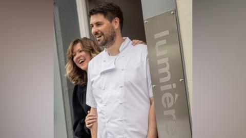 A husband and wife team smile as they stand in the doorway to their restaurant, Lumiere. The woman has mid-length dark blonde hair and is smiling as she stands behind her husband and rests her hands on his shoulder and arm. He has dark hair and stubble, is wearing chefs whites and is smiling.