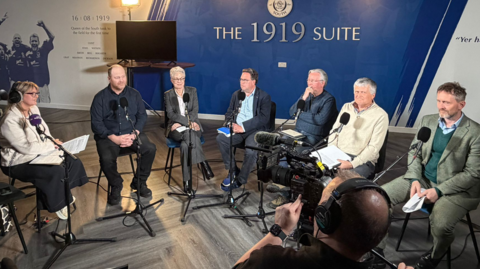 Group of five men and two women in a room. They are talking into large microphones.