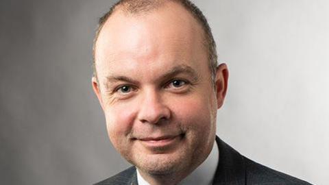 A white man wearing a suit. It is a professional photograph. He is against a plain grey background. He is smiling at the camera. His hair is receding slightly. He is clean shaven. 