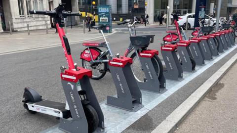 Red and white e-scooters and e-bikes at a docking bay in the city. They have LNER branding on the sides of them.