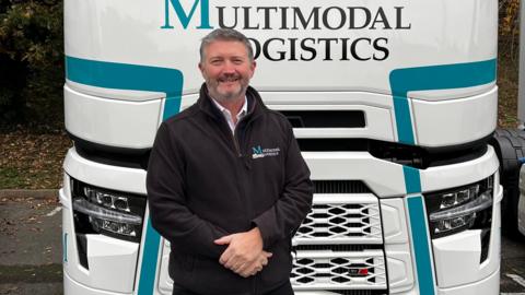 Tim Wray smiles at the camera in front of a white HGV lorry. He has short grey hair and wears a black jumper with his hands resting in one another in front of him.