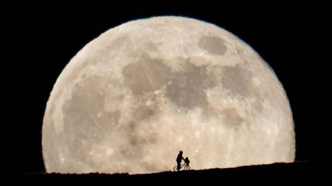 Silhouette of boyish character in an anorak straddling a stationary BMX bike with an object in its front basket on a mountainside framed by the full Moon behind