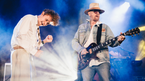 Tom A. Smith is smiling as he shakes his head in the air next to Dave Stuart, who is playing the guitar. Smith is wearing beige trousers with a brown belt, a brown and beige striped tie and white shirt. Stuart is wearing jeans and a denim jacket over a white shirt. He has tattoos on his neck and fingers and a beige hat and sunglasses. 
