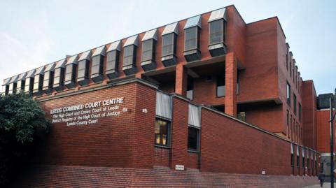 A bricked building with several windows. There is white writing on the building which says "Leeds Combined Court Centre The High Court and Crown Court at Leeds District Registry of the High Court of Justice Leeds County Court".