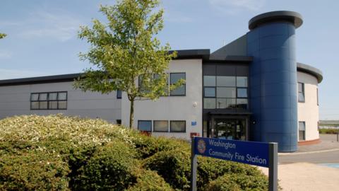 The two storey modern building is made of various blue, white and brown materials. There is a tall blue column running up the middle. The building is set in from the main road, behind a pavement area with hedges and a tree on it. A sign in the foreground reads: "Washington Community Fire Station."