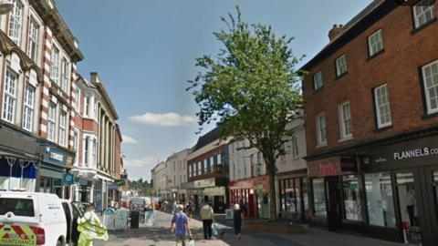 A Google view of Commercial Street in Hereford. There are shops on both side of the pedestrianised street, which is seen in the day time. Shoppers are walking around with bags and a contractor is standing near a van.