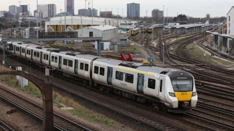 A Thameslink train on the tracks passing through Selhurst in London with a low railway shed building in the background and beyond it tower blocks 