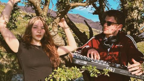 Ruby ar y chwith ac Endaf ar y dde yn sefyll mas tu fas wrth ymyl coeden. Mae gan Ruby wallt hir brown ac mae'n gwisgo crys-t du. Mae Endaf ar y dde yn gwisgo yn gwisgo clust-ffonau ac yn dal bysellfwrdd