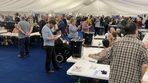 A tent erected inside a sports arena. A blue carpet has been laid down and spotlights illuminate the space. Tables are lined up with people counting votes.
