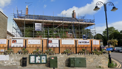 A red brick two-storey house with a chimney on either side. The building is entirely covered in scaffolding, with a construction fence and a black gate blocking the view of the ground floor.