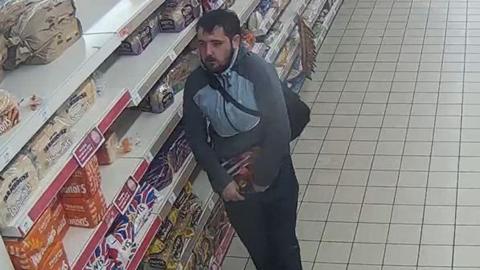 A man in a grey tracksuit stood in a aisle of a supermarket holding a packet in one hand and pulling out the waist of his trouser with the other.