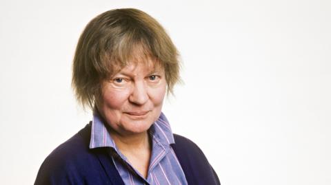 Dame Iris Murdoch, with short hair above the shoulder, staring into the camera wearing a striped purple shirt with upturned collar and purple cardigan