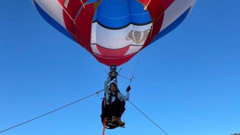 Jo Bailey is pictured in a seat attached to a hot air balloon, against a cloudless blue sky. The balloon is red, blue and white; Bailey has dark hair and wears a light blue top, black trousers and brown boots. 