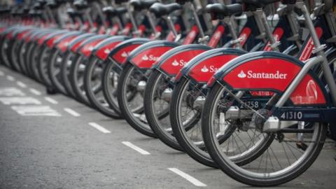 A row of Santander Cycles bicycles parked along a road in a designated bay, marked by white dashed lines. The back wheels of the bicycles are visible, with a red partial wheel cover displaying the Santander logo.