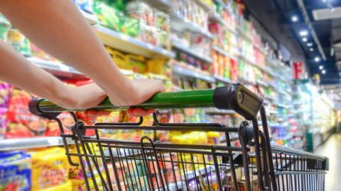 A shopping trolley being pushed in a supermarket. Hands visible