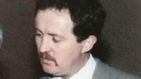 A PSNI handout image of John Devine. He has dark hair and a moustache. He is wearing a grey suit and white shire. 