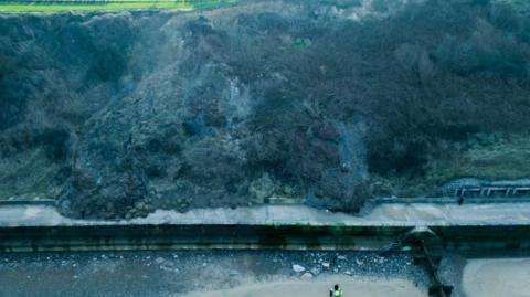 A cliff-fall shows debris on a path and barriers and people in hi-vis jackets standing in the sea at the foot of a cliff