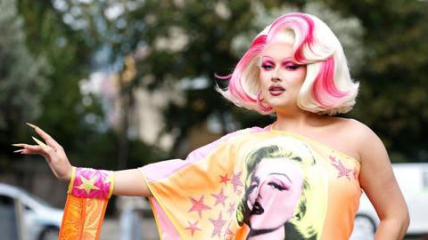 Elle Vosque is wearing a blonde wig with pink highlights. She is wearing an orange off the shoulder dress decorated with pink stars and a picture of Marilyn Monroe.