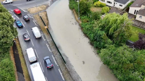 A view of a road approaching a roundabout with traffic on the left handside. The right hand side of the road is covered in water. 