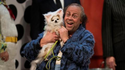 Baritone David Stout clutches Bridget the dog - dressed in a cat costume - in the closing scene from Don Pasquale. He is wearing a tatty blue coat and is grinning with happiness.