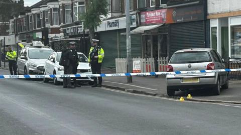 A silver VW Polo on the pavement in front of a row of shops. There is a police cordon in place, with tape across the road.