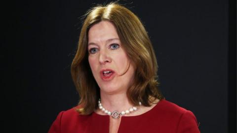 Dr Catherine Calderwood, with brown hair and wearing a red top and white pearl necklace, speaks in front of a dark background at a press conference 