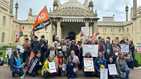 A group of workers hold banners and signs outside an Indo-Islamic-style building. 