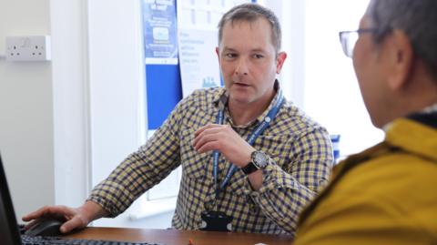 A male Citizens Advice volunteer wearing a blue and yellow chequered shirt sat at a desk with a computer on it talks to a person sat opposite him.