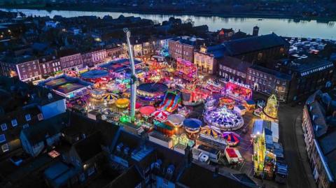 A view from a drone of King's Lynn's town centre with the Mart set up on the Tuesday Market Place. Its rides are lit up in a rainbow of colours. Behind the town is the river Great Ouse and a beautiful orange sky. 