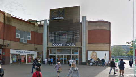 A Google Street image of the outside of the County Mall in Crawley. You see a number of people walking outside the entrance which has a sign which says "COUNTY MALL" above the sliding doors. At the top there is a large "CM" with "COUNTY MALL" below it.