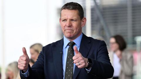 Rhun ap Iorwerth, a man with short brown hair in a navy suit, with his thumbs up.