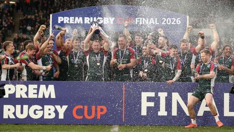 Leicester players hold the trophy in the air and spray champagne while standing in front of the winners' podium