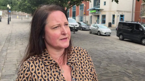 Close up of Alison Hernandez. She has dark hair and is wearing a beige and black blouse. She is looking to the side of the camera. Ms Hernandez is stood on a street, cars are parked behind her.