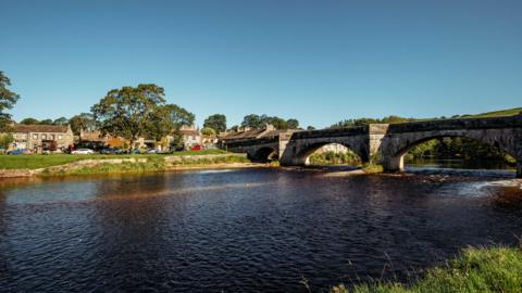 Three arches of a stone bridge can be seen spanning the River Wharfe on a sunny day. The stone houses of the village can be seen on the far side of the river.