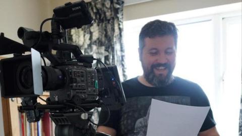 A man smiling while standing next to a black camera. He has a long beard, and is holding a white piece of paper, which he is looking at.