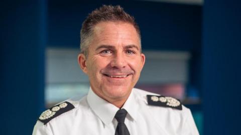 A Simon Tuhill pictured smiling with short dark spikey hair. He is wearing a white shirt and black tie with black Epaulettes on his shoulders.