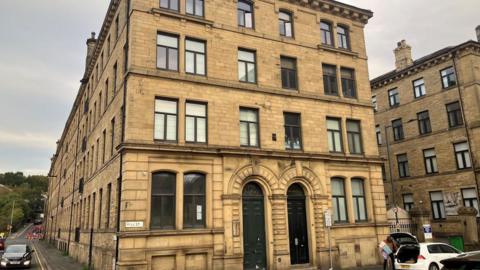 A large, four-story building constructed from light brown stone. Each floor has several rectangular windows framed in a dark material. On the ground floor, there are two prominent arched doorways fitted with dark green doors. A street sign on the corner of the building reads "MILL ST."
In front of the building, a cobblestone street runs along the edge, where several cars are parked. One white car has its boot open and a person is standing beside it. The sky is overcast. Adjacent to the main building, another similar stone structure is visible on the right.