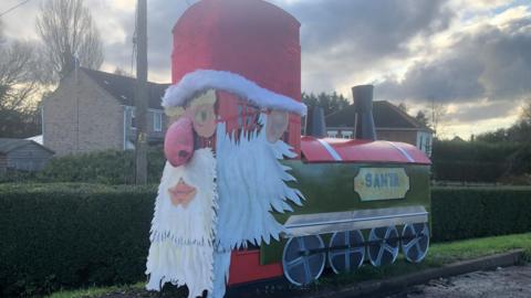 A Santa train on phone box. A giant Santa face including beard, a hat and a large red nose covers the telephone box while the train engine, which is green and red is made from plywood and aluminium - with plant pots for the chimneys