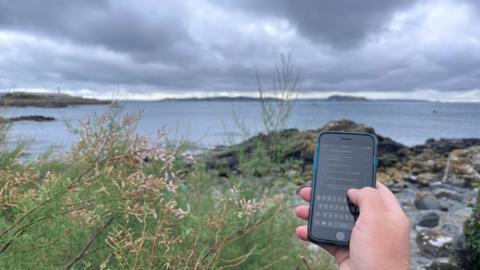 A hand holds a phone with a search for Guernsey Broadband on the screen, in front of a beach and some shrubbery, as well as islands in the distance.