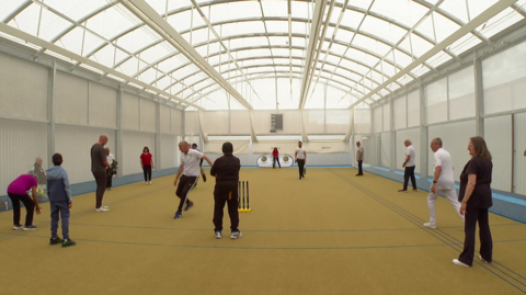 A large sports hall with white walls, an arched roof and yellow floor with people standing around and cricket stumps in the foreground
